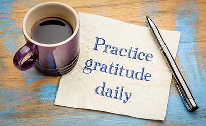 Coffee cup on a desk next to a napkin and pen. Practice Gratitude Daily is written on the napkin in blue ink.