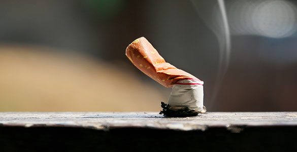 A cigarette has been put out on a flat stone table or bench. It is still smoking. There is a blurred, green, outdoor type of background