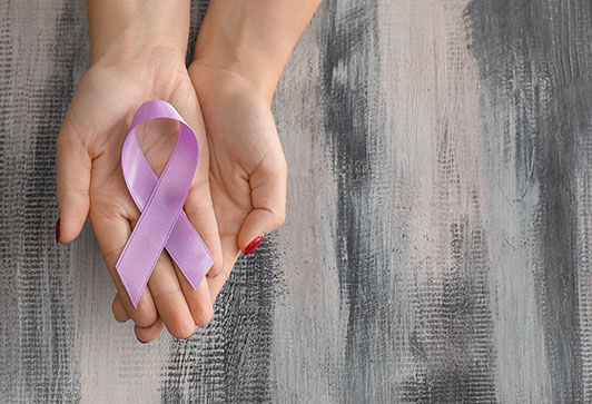 Two overlapping hands palms up and open holding a lavendar awareness ribbon on top of a gray weathered wood table