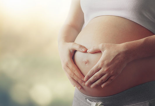 Image of a woman's pregnany belly, her own hands placed on her stomach outlining the belly button in a heart shape