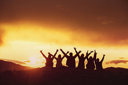 Sunset in the background, six people sit on top of a hill with their arms raised