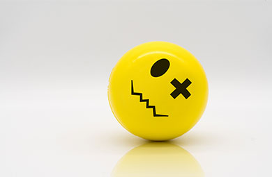 Yellow stress ball an x for one eye. The mouth is sqwiggly with hard angles.